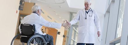 Two doctors shaking hands in hospital corridor, one with spinal cord injury and in a wheelchair.