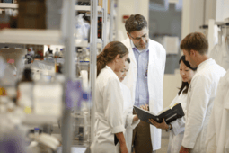 Five scientists in lab coats review a notebook together in a laboratory setting, surrounded by equipment and supplies.
